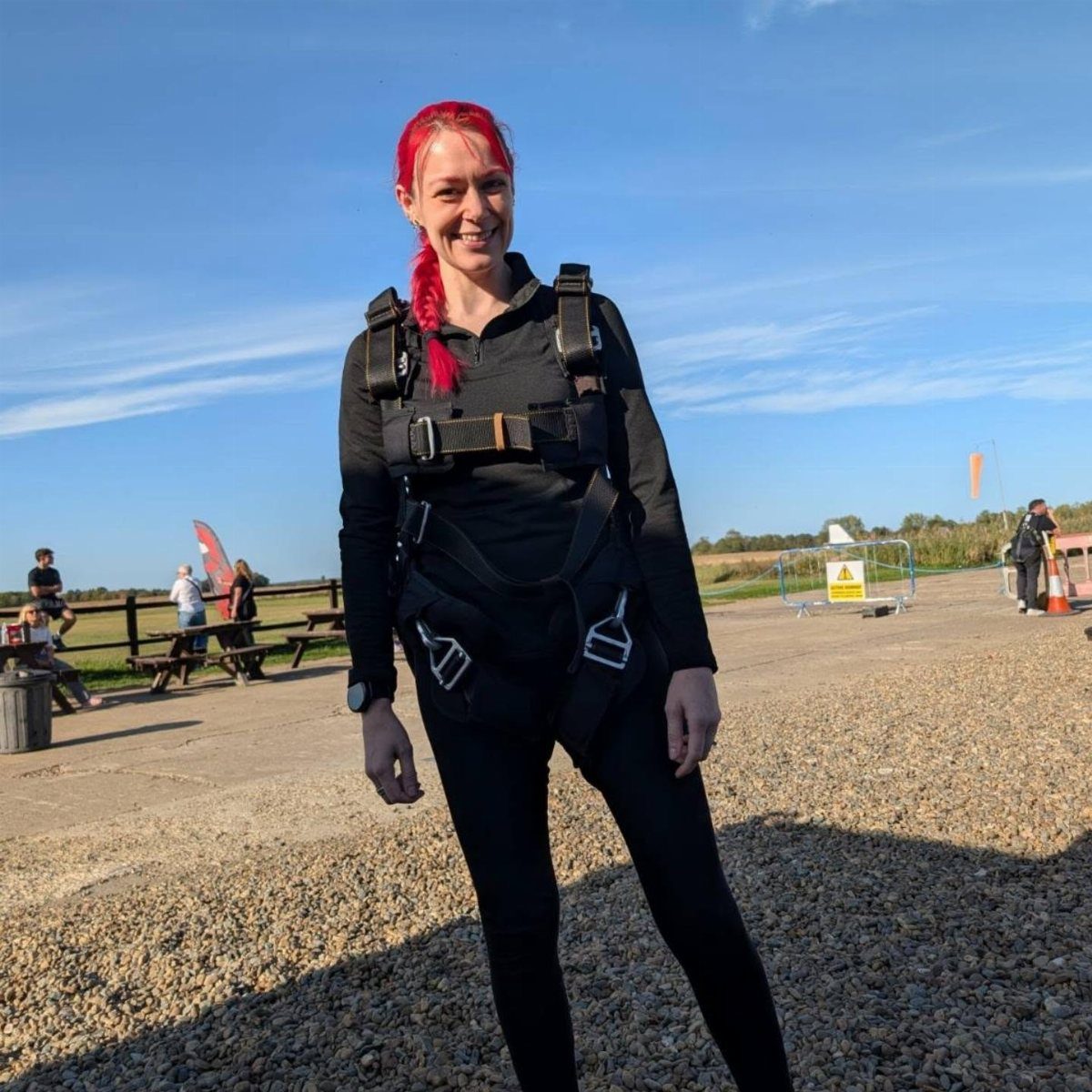 Woman with red hair wearing a skydiving harness standing on a gravel airfield.