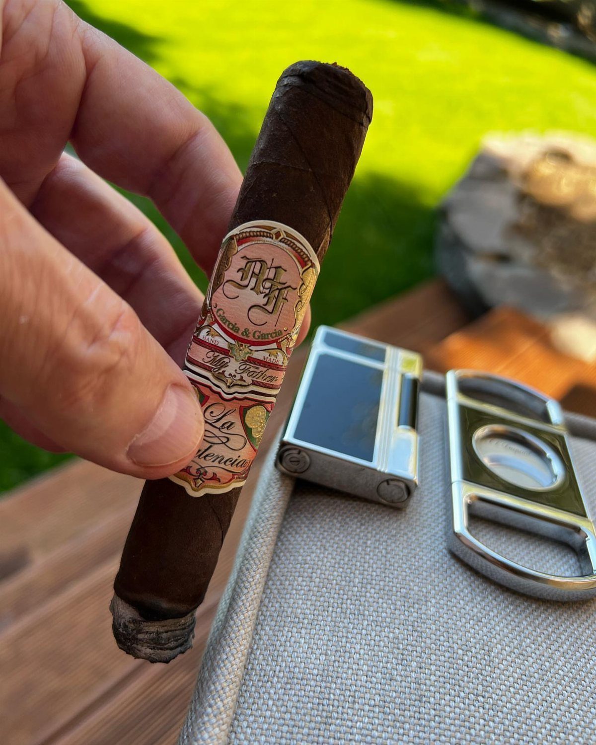 Hand holding a brown cigar next to a silver cutter and lighter outdoors.